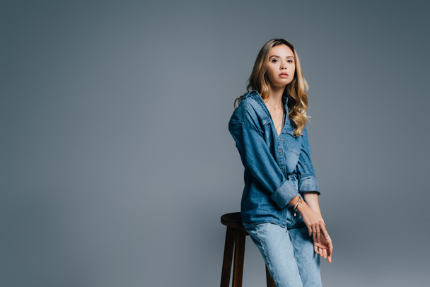 stylish woman in denim clothes near high stool isolated on grey - Foto, afbeelding