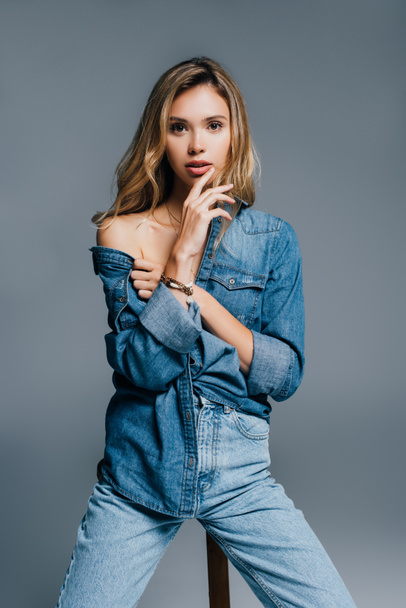 sexy young woman in denim shirt touching lips while posing with naked shoulder isolated on grey - Photo, Image
