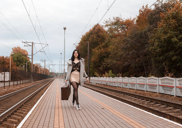 молодая улыбающаяся девушка в сером пальто с чемоданом, идущая вдоль железнодорожного вокзала осенью - Фото, изображение