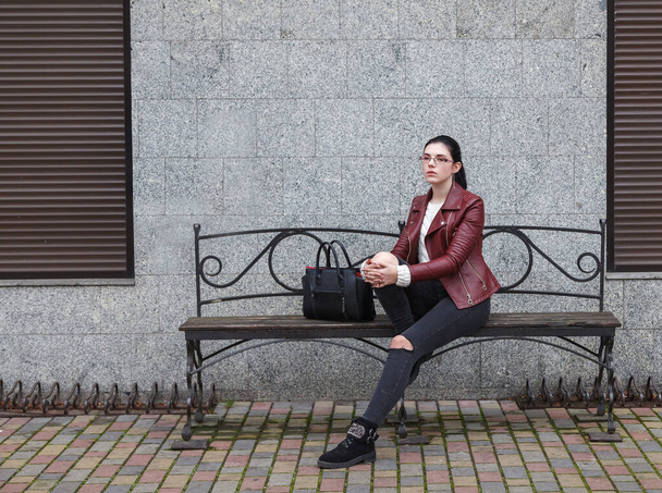 solitario giovane ragazza in una giacca marrone e jeans neri seduti su una panchina vicino al marciapiede nella cupa giornata autunnale - Foto, immagini