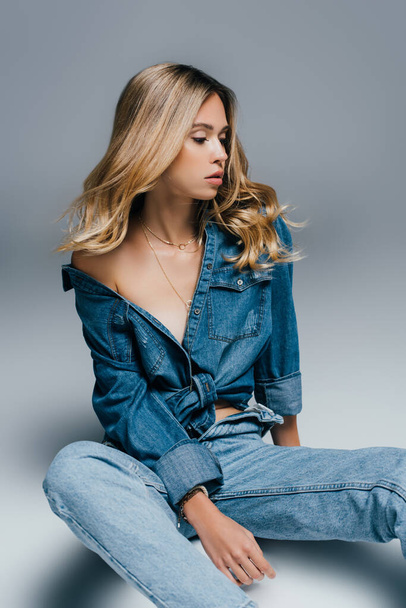 чувственная молодая женщина в расстегнутой джинсовой одежде сидит на сером - Фото, изображение