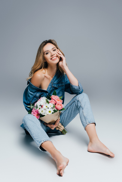 cheerful barefoot woman in denim clothes sitting with bouquet on grey - Foto, Bild