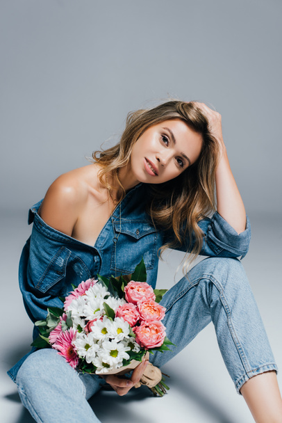sensual young woman in denim clothes touching hair while sitting with flowers on grey - Foto, Imagem