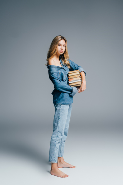 sexy barefoot woman in denim clothes, with naked shoulder, holding stack of books on grey - Fotoğraf, Görsel