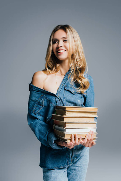 young seductive woman in denim shirt, with naked shoulder, holding books isolated on grey - Fotoğraf, Görsel
