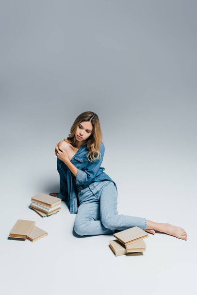 young sensual woman in denim clothes touching naked shoulder while sitting near books on grey - Fotografie, Obrázek