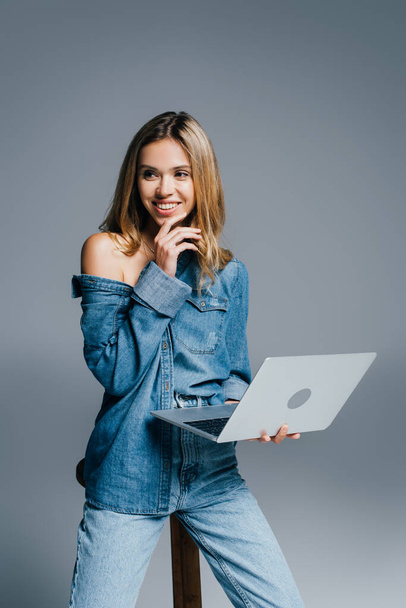 smiling woman in denim shirt and jeans looking away while holding laptop on grey - Fotoğraf, Görsel