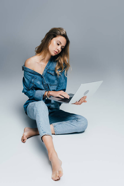barefoot woman in denim shirt and jeans using laptop while sitting on grey - Fotó, kép