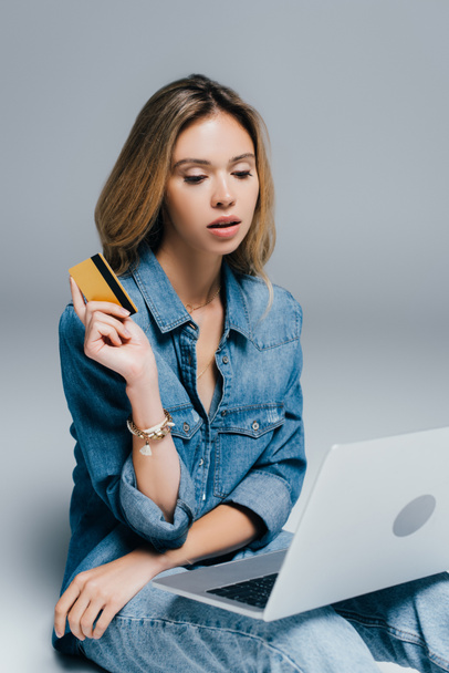 stylish woman in denim clothes holding credit card while looking at laptop on grey - Фото, изображение