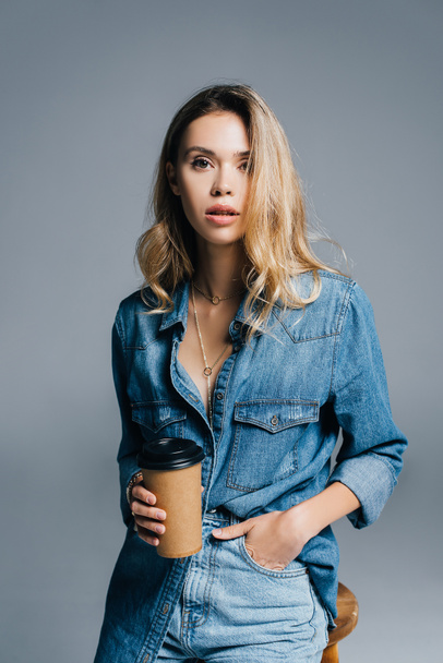 stijlvolle vrouw in denim kort, met de hand in zak, het houden van koffie te gaan geïsoleerd op grijs - Foto, afbeelding