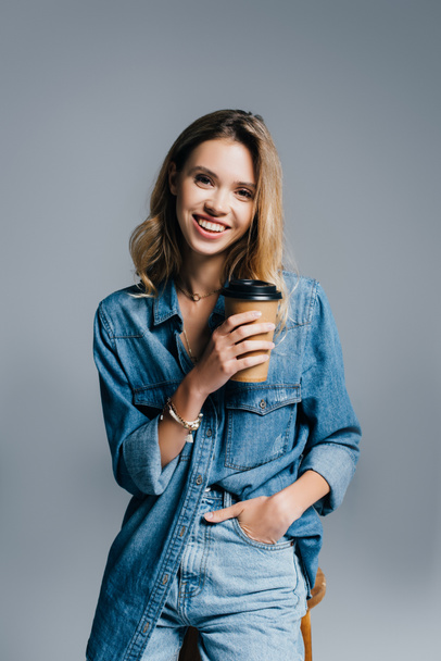 uśmiechnięta młoda kobieta w dżinsowych ciuchach trzymająca rękę w kieszeni i kawę, by odizolować się na szaro - Zdjęcie, obraz