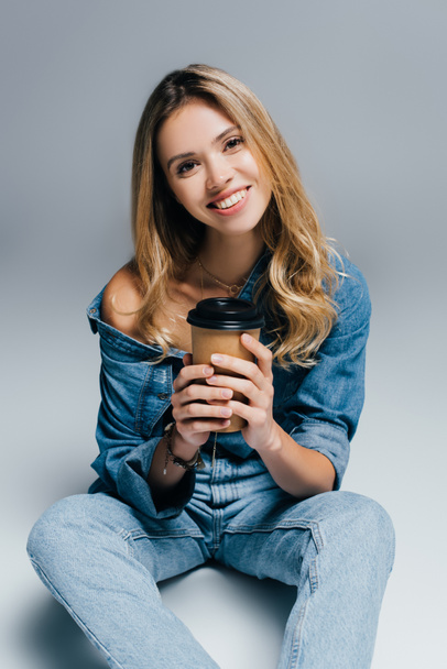 happy young woman in denim clothes, with naked shoulder, holding coffee to go while sitting on grey - Fotó, kép
