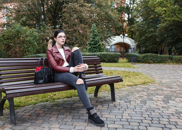 νεαρή κοπέλα με καφέ σακάκι και μαύρο τζιν κάθεται σε ένα παγκάκι πάρκο το φθινόπωρο - Φωτογραφία, εικόνα