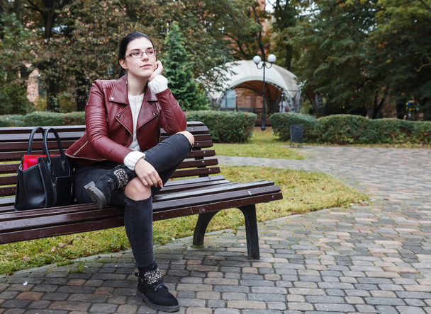 jong meisje in bruine jas en zwarte jeans zitten op een park bank in de herfst - Foto, afbeelding