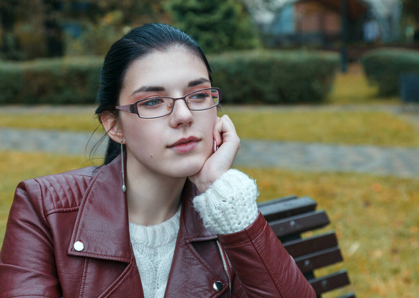 Осенью молодая девушка в коричневой куртке и черных джинсах сидит на скамейке в парке. портрет крупным планом - Фото, изображение