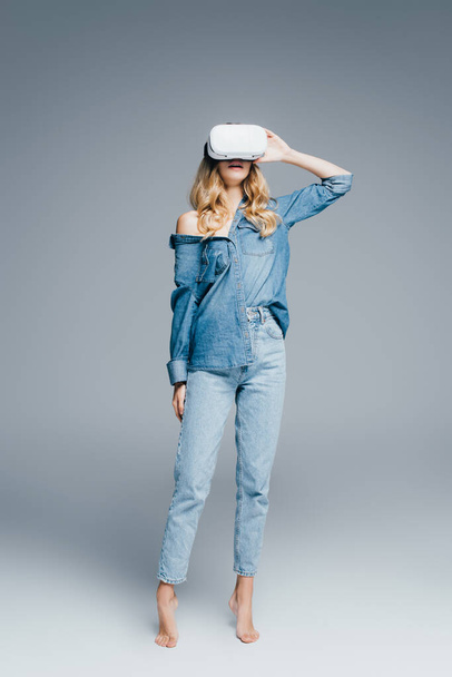 vue pleine longueur de femme sensuelle en jeans et chemise en denim en utilisant un casque vr sur fond gris - Photo, image