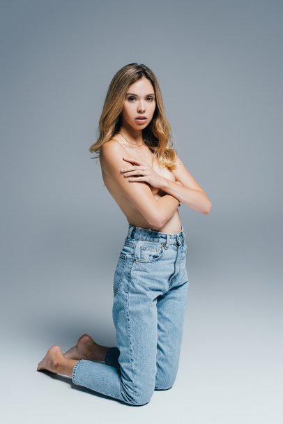 topless, seductive woman in jeans hiding breasts with hands while kneeling on grey - Фото, зображення