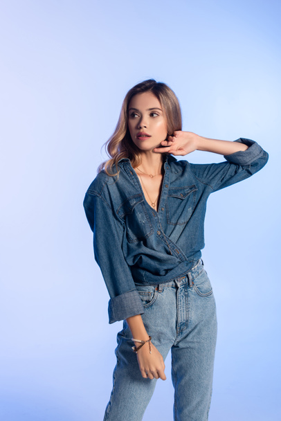 young woman in denim shirt and jeans touching face while looking away isolated on blue - Photo, image