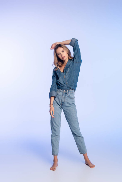 vista completa de la joven mujer descalza en camisa de mezclilla y jeans posando en azul - Foto, Imagen