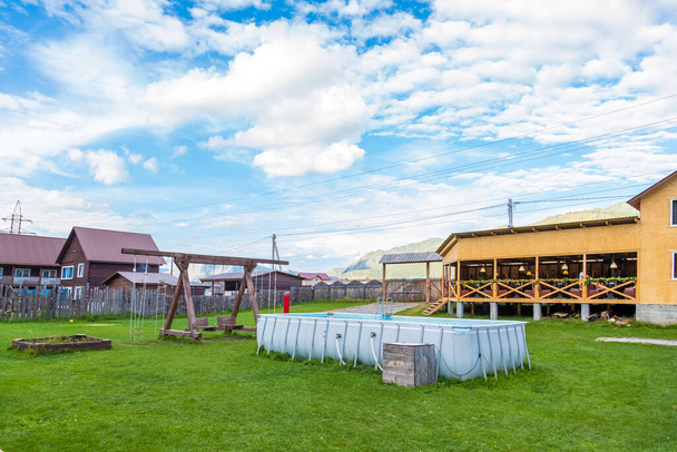 Une grande balançoire en bois et une piscine gonflable en caoutchouc sur une pelouse d'herbe verte dans la cour d'une maison de campagne avec une clôture en bois sur fond de montagne et de ciel bleu. - Photo, image