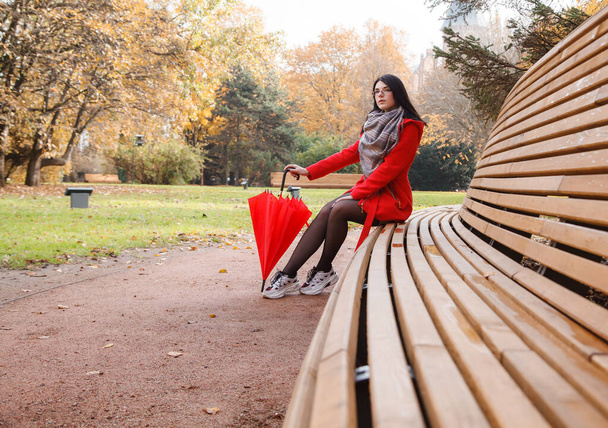 young girl in a red coat with an umbrella sitting on a bench in a city park after the rain on an autumn day - Photo, image