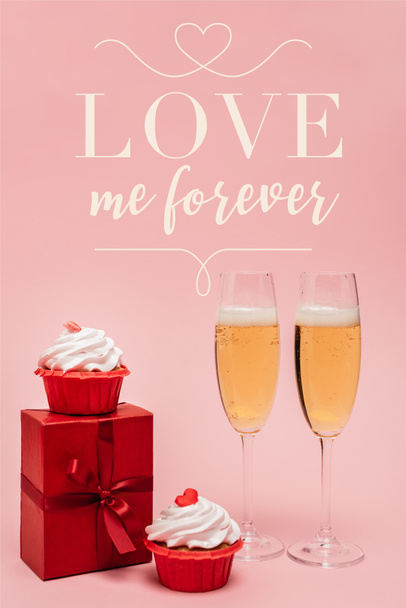 champagne glasses near gift box, cupcakes and love me forever lettering on pink - Photo, image