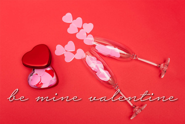 Top view of pink hearts in champagne glasses near metallic box and be mine valentine lettering on red background - Фото, изображение
