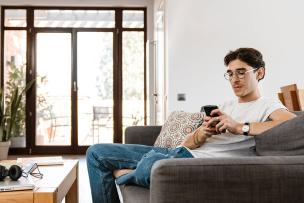 Image of concentrated young handsome man sitting on sofa indoors at home while using mobile phone - Foto, Imagem