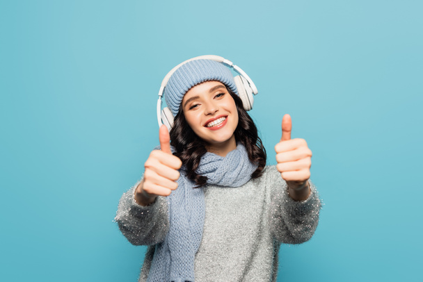 Boldog nő kalap és sál hallgat zenét fülhallgató és mutatja hüvelykujját elszigetelt kék - Fotó, kép