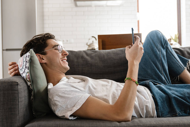 Picture of optimistic pleased handsome man lies on sofa indoors at home using mobile phone - Fotografie, Obrázek