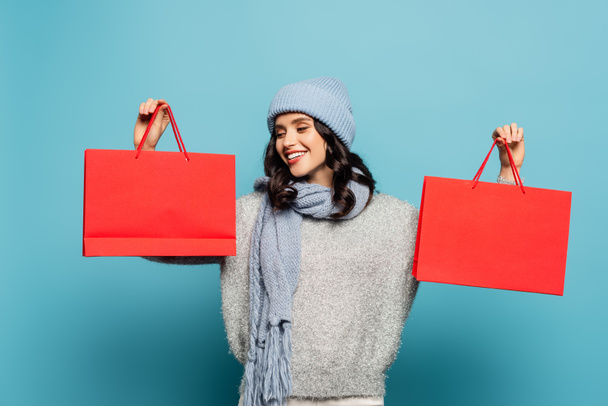 Cheerful brunette woman wearing winter outfit and looking at red shopping bag isolated on blue - Valokuva, kuva
