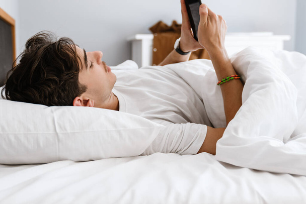 Image of handsome man lies in bed and using mobile phone indoors at home - Foto, afbeelding