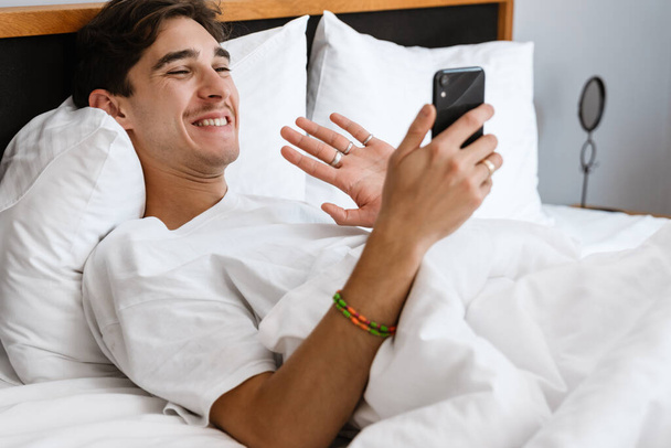 Image of handsome cheerful man lies in bed and talking by videocall by mobile phone indoors at home while waving - Photo, Image