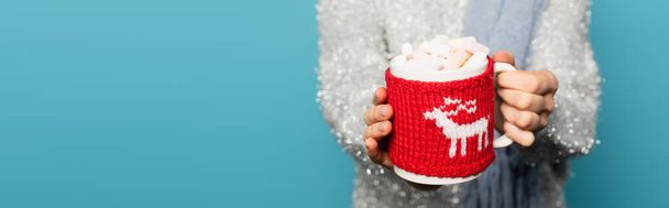 Close up view of cup with marshmallows and knitted holder in hands of blurred woman on background isolated on blue, banner - Photo, Image