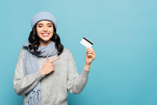 Happy brunette woman in winter outfit pointing with finger at credit card while looking at camera isolated on blue - Фото, изображение