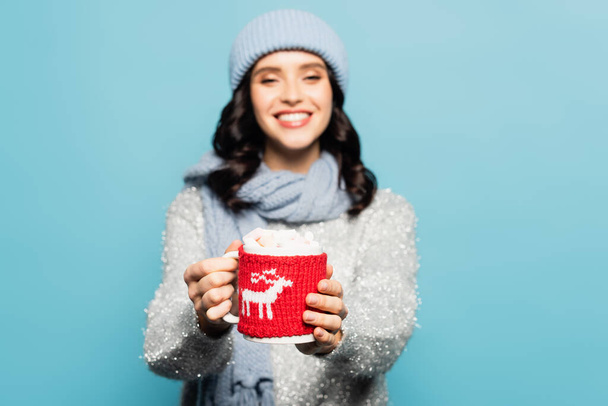 Cup with marshmallows and knitted holder in hands of blurred woman on background isolated on blue - Foto, Bild