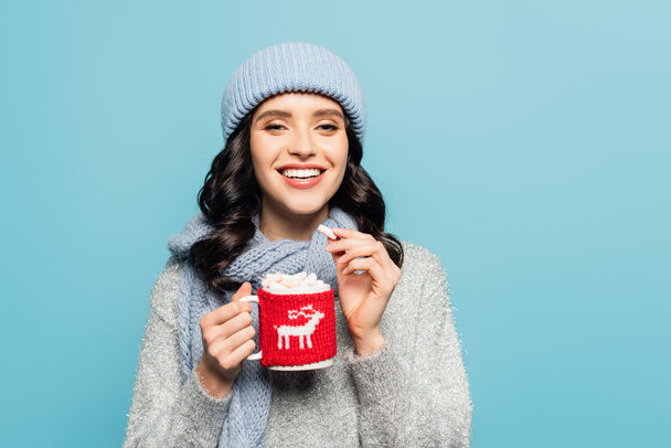 Front view of happy woman in hat and scarf holding cup and marshmallow while looking at camera isolated on blue - Fotografie, Obrázek