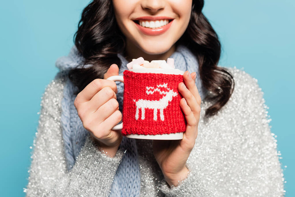 Cropped view of brunette woman in scarf holding cup with marshmallows and knitted holder isolated on blue - Fotografie, Obrázek