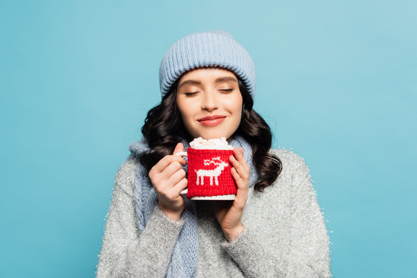 Smiling brunette woman in winter outfit, with closed eyes holding cup with marshmallows isolated on blue - Zdjęcie, obraz