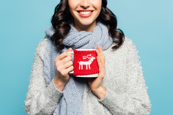 Cropped view of brunette woman in scarf and pullover holding cup with knitted holder isolated on blue - Photo, Image
