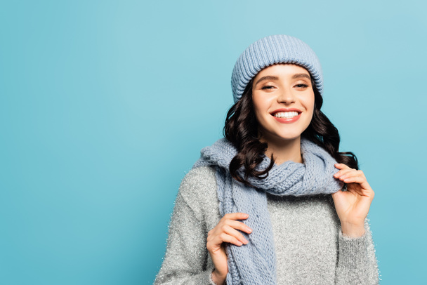 Happy brunette woman in winter outfit touching scarf while looking at camera isolated on blue - 写真・画像