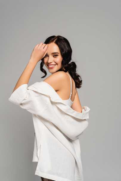 Happy brunette woman in white top and shirt looking at camera while posing isolated on grey - Foto, afbeelding