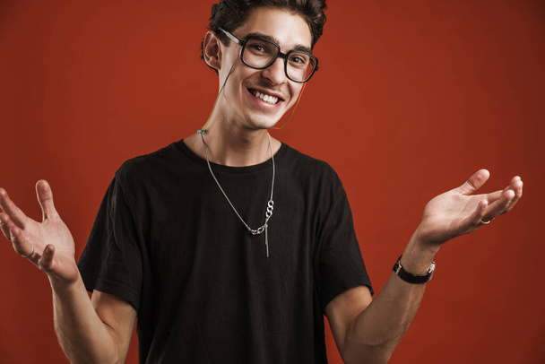 Confused smiling young hipster man standing isolated over red background, shrugging shoulders - Foto, Bild
