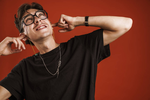 Close up of a young teenage boy wearing glasses standing isolated over red background, covering ears - Foto, Imagen