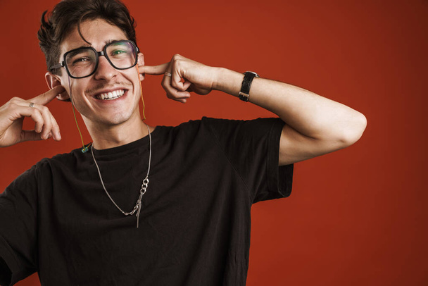 Close up of a smiling young teenage boy wearing glasses standing isolated over red background, covering ears - Foto, Bild