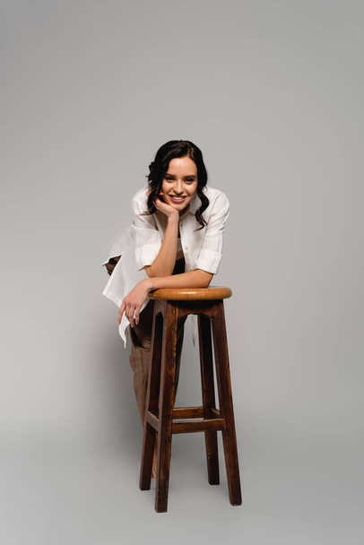 Full length of smiling brunette woman in white shirt looking at camera while leaning on stool on grey - Photo, Image