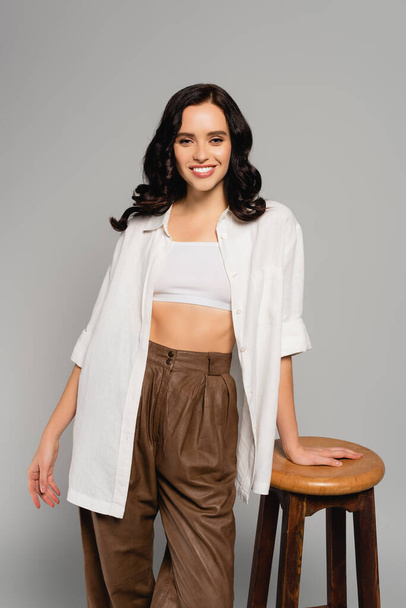 Smiling brunette woman in white top and shirt looking at camera while leaning on stool isolated on grey - Foto, Imagem