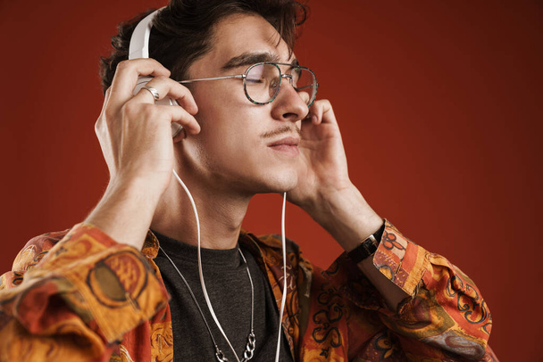 Young man enjoying music, listening through wireless headphones isolated on red background - Foto, imagen