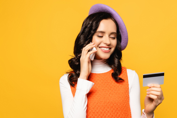 Smiling brunette woman in beret talking on cellphone while looking at credit card isolated on yellow - Фото, зображення