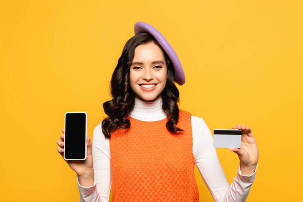 Positive brünette Frau mit Baskenmütze blickt in die Kamera und zeigt Kreditkarte und Smartphone isoliert auf gelb - Foto, Bild